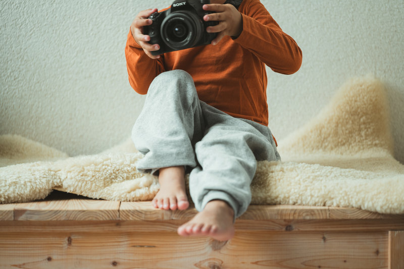 A child in an orange top holding a camera, sitting barefoot on a wooden bench